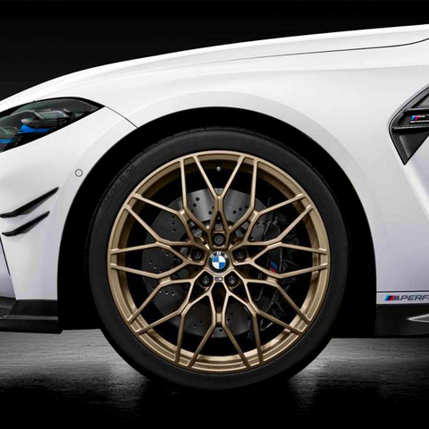 A gold multi-spoke alloy wheel with the BMW logo is attached to a white car, featuring black sports detailing and "M Performance" decal, set against a dark background.