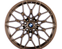 Bronze car wheel with intricate, multi-spoke design, centered on a logo featuring black, white, and blue colors, resting against a plain white background.
