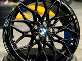 A glossy black BMW wheel rests against stacked tires in a garage. Its intricate spokes and central logo are prominent with a blurred background of tires and tools.