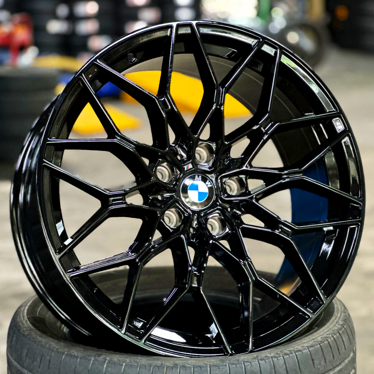 A shiny, black BMW wheel rests upright on a tire, showcasing intricate, angular spokes; set within an automotive workshop environment with blurred stacks of tires and tools in the background.
