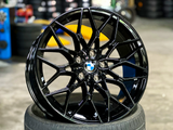 Black, multi-spoke alloy wheel with a BMW logo, resting upright on a tire. The background shows a garage setting with stacked tires and equipment.