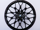 A black alloy wheel with a complex, geometric spoke design sits against a plain white background. At the center is a BMW logo, featuring blue, white, and black colors.