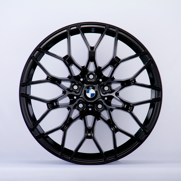 A black, intricately designed car wheel rim with a central blue and white logo is displayed against a plain white background.