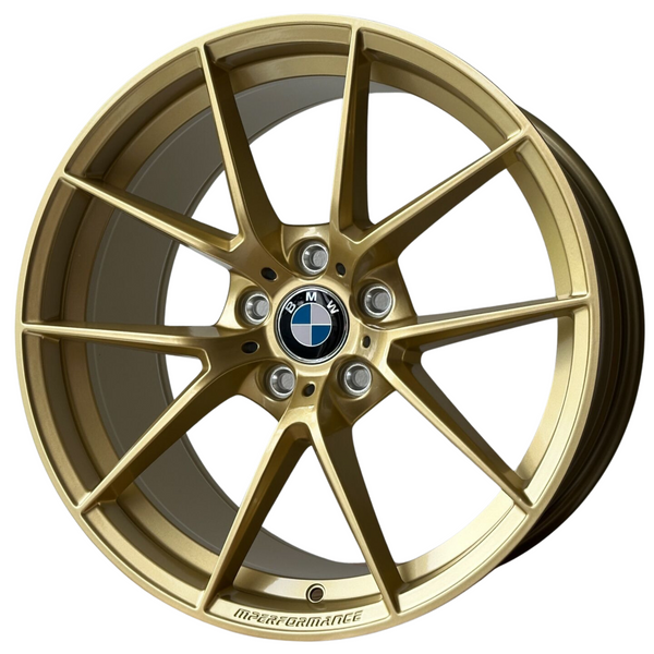 A gold alloy wheel with a central BMW logo and "M PERFORMANCE" text is isolated on a white background, showcasing intricate spokes and bolt holes.