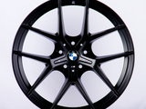 A black BMW wheel rim with a multi-spoke design is displayed upright against a plain white background. The BMW logo is centered, and the 