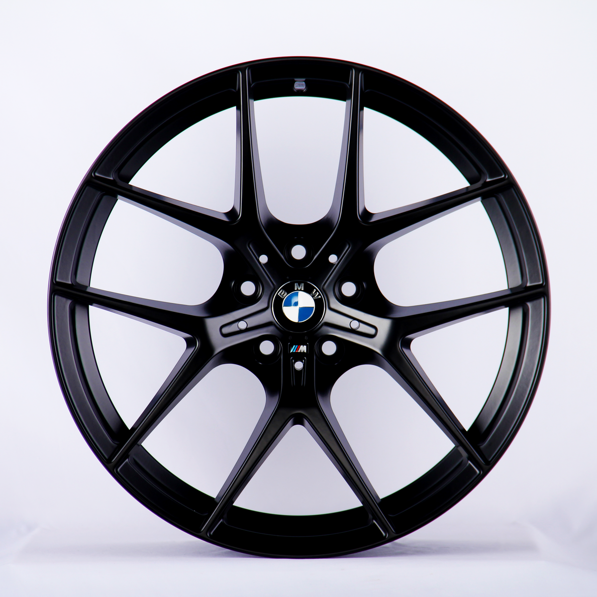 A black BMW wheel rim with a multi-spoke design is displayed upright against a plain white background. The BMW logo is centered, and the "M" insignia appears below it.
