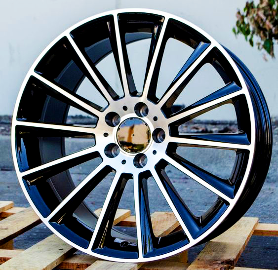 A shiny, black multi-spoke alloy wheel stands upright on a wooden pallet, situated outdoors on a concrete surface with a white wall and some greenery in the background.