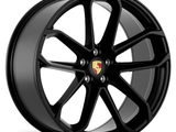 A black alloy wheel with a five double-spoke design, featuring the Porsche crest at its center, is positioned on a white background.