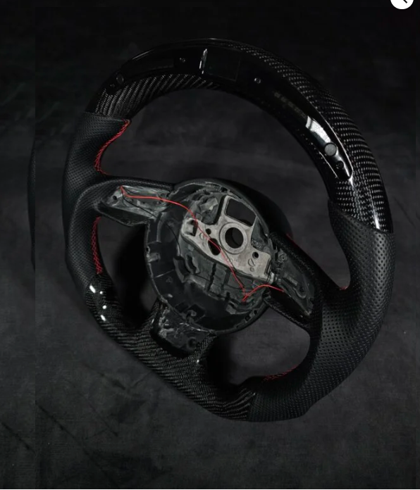 A black carbon fiber steering wheel with red stitching, shown without a center cover, is positioned on a dark, shadowy background.