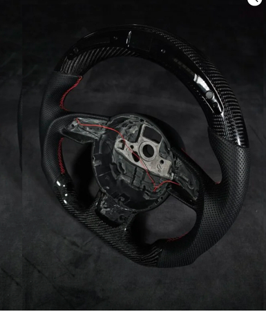 A black carbon fiber car steering wheel with red stitching, partially assembled, sits on a dark surface. It has visible wiring and an exposed central mounting area.