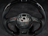 A carbon fiber steering wheel with missing center components and exposed red wires lies on a dark surface, highlighting its textured grips and sleek design.