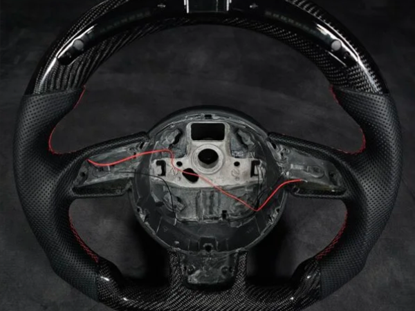 A carbon-fiber steering wheel missing its center, with exposed wiring, rests against a dark background, highlighting red stitching accents on the grips.