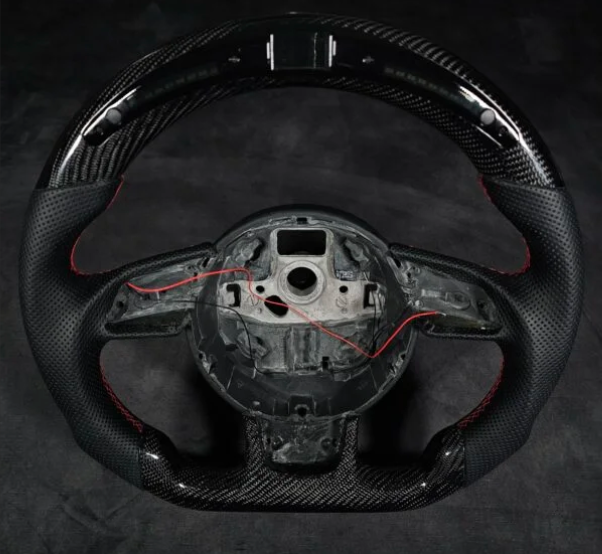 A sleek, carbon fiber car steering wheel with red stitching shows exposed internal wiring and mounting points, set against a plain dark background.