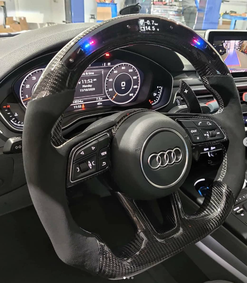 A carbon-fiber steering wheel with LED indicators is central, featuring control buttons and the Audi logo. It's in a car interior with a digital dashboard displaying "11:52 AM," 11/16/2020."