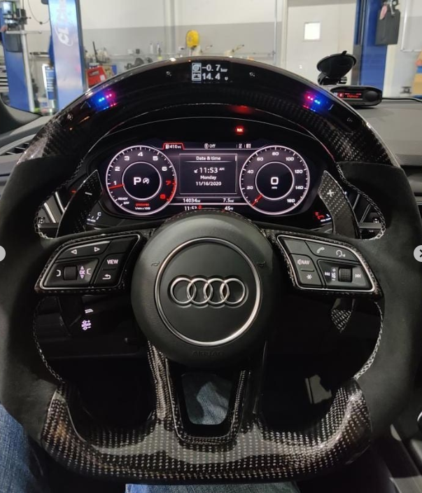 A carbon fiber steering wheel with controls, displaying a digital dashboard showing speed and information, inside an Audi car, in a workshop setting.