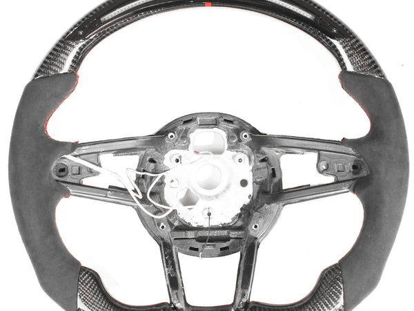 Steering wheel, disassembled, showing a sleek design with carbon fiber and suede accents; some internal wiring is exposed, against a plain white background.