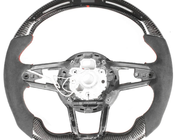 Steering wheel, disassembled, showing a sleek design with carbon fiber and suede accents; some internal wiring is exposed, against a plain white background.