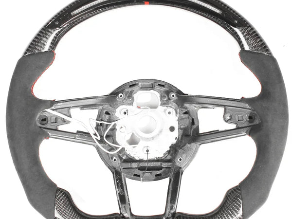 The object is a racing-style car steering wheel with carbon fiber and suede trim. The central section is unfinished, showing wiring and mounting points, against a plain white background.