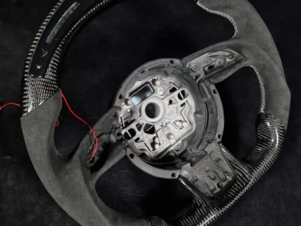 A carbon fiber steering wheel with exposed wiring lies on a dark surface, featuring a central mounting area and a mix of textured and smooth materials surrounding the wheel.
