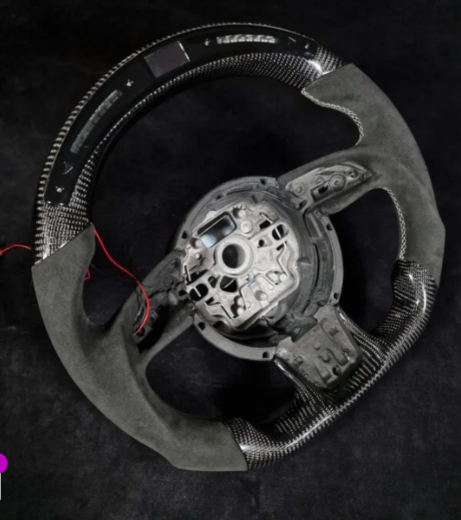 A car steering wheel, partially disassembled, with carbon fiber and suede elements. Red wires are exposed, resting on a dark surface.