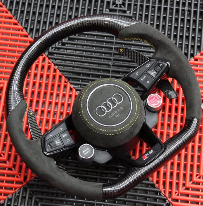 Steering wheel featuring Audi logo and controls, placed on a textured red and black surface. Notable buttons include "view," "nav/map," "drive select," and a red "RS" label.
