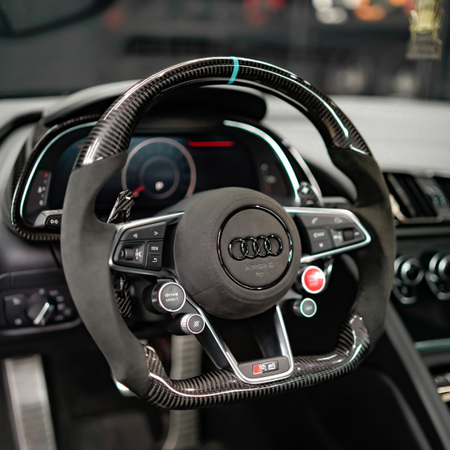 A steering wheel with carbon fiber accents features various control buttons, including "drive select" and "view," set within a modern Audi dashboard displaying digital gauges in a sleek car interior.