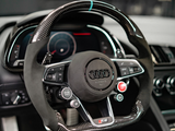 Steering wheel featuring carbon fiber and leather design, adorned with the Audi logo, surrounded by dashboard controls and digital displays within a car's interior.