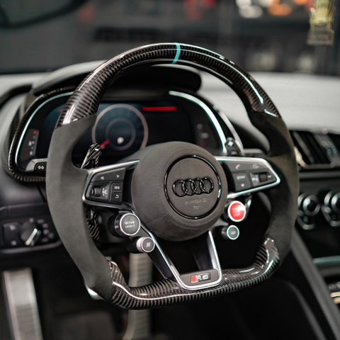 Steering wheel featuring carbon fiber and leather design, adorned with the Audi logo, surrounded by dashboard controls and digital displays within a car's interior.
