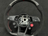 A carbon fiber steering wheel with black and red accents is displayed against a dark background. It features several buttons including 
