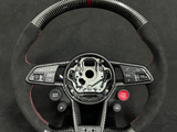 A carbon fiber steering wheel, featuring control buttons and a red stripe at the top, is set against a dark background. Notable texts include 
