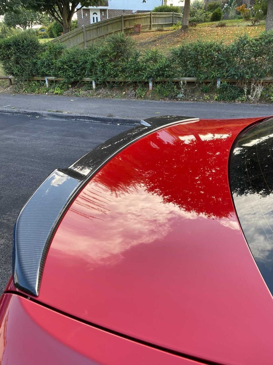 A red car with a carbon fiber spoiler reflects clouds, parked near an asphalt road. The background features green shrubs, a sloped wooden fence, and trees.