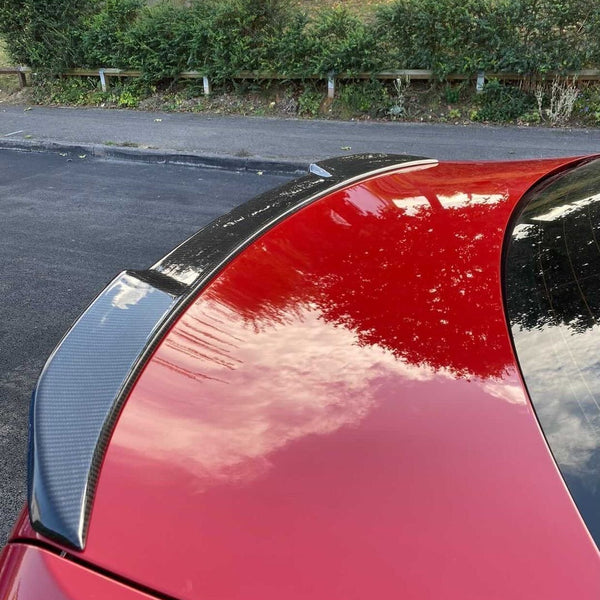 A red car with a carbon fiber spoiler reflects clouds, parked near an asphalt road. The background features green shrubs, a sloped wooden fence, and trees.
