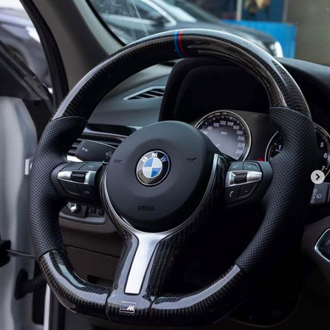 A carbon-fiber steering wheel with a BMW logo is centered in a car's interior, surrounded by a dashboard and speedometer. The text reads: "BMW" and "AIRBAG."