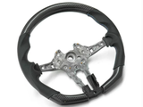 A black steering wheel with metal framework, featuring blue stitching and carbon fiber-like texture, rests on a plain white background.