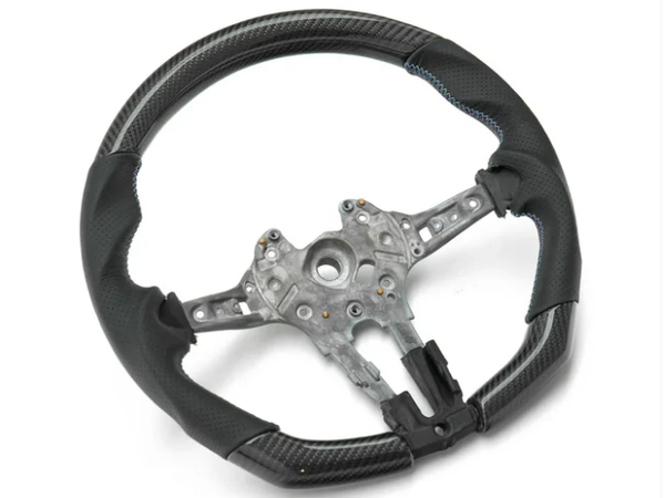 A black steering wheel with metal framework, featuring blue stitching and carbon fiber-like texture, rests on a plain white background.