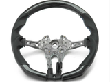 A partially assembled car steering wheel is shown, featuring a metallic central frame with exposed wiring, surrounded by a two-tone black and grey perforated leather grip, against a plain white background.