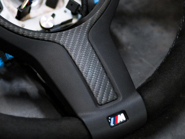 Steering wheel, featuring a carbon fiber insert and "M" logo, rests on a dark surface, with exposed wiring visible in the center.