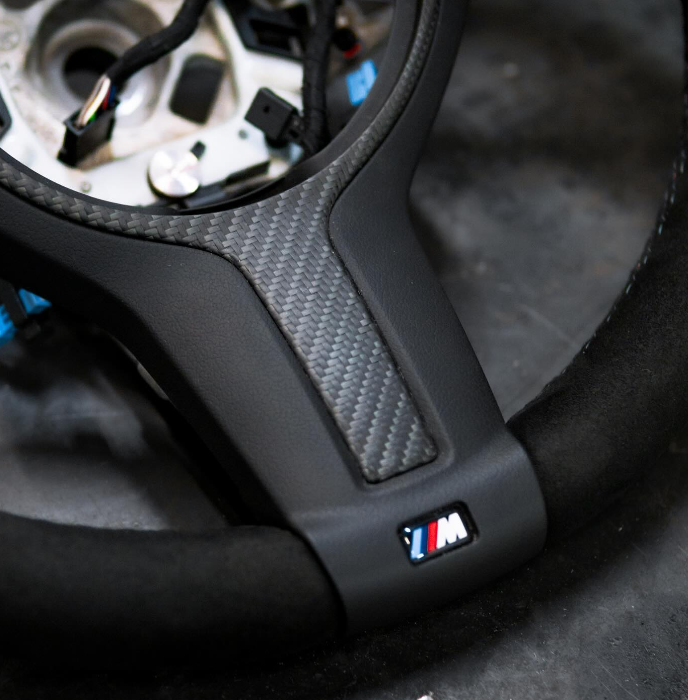 Steering wheel, featuring a carbon fiber insert and "M" logo, rests on a dark surface, with exposed wiring visible in the center.