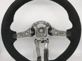 A circular car steering wheel with a visible metal framework is standing upright. It lacks a central cover, revealing internal fittings, and rests against a plain white background.