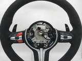 A black suede car steering wheel with a red top stripe is shown, featuring paddle shifters and multiple control buttons, set against a plain white background. At the bottom, there's an 