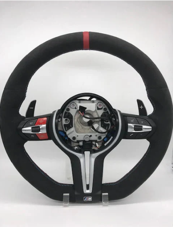 A black suede car steering wheel with a red top stripe is shown, featuring paddle shifters and multiple control buttons, set against a plain white background. At the bottom, there's an "M" emblem.