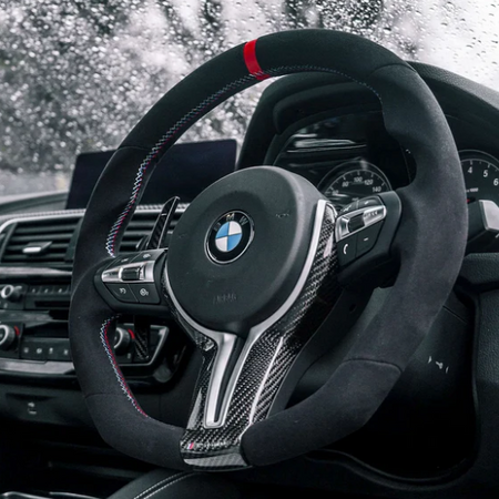 Steering wheel with BMW logo features a sleek design and controls. It's set against a car dashboard with raindrops visible on the windshield, implying a rainy day outside.