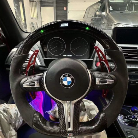 A BMW steering wheel with red paddle shifters is centered, displaying the logo and "M" branding. The dashboard shows speed dials. Interior features include camouflage-patterned pants and a parked car outside.
