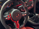 The steering wheel, featuring carbon fiber detailing and control buttons, is stationary in a car interior with red accents, digital dashboard, and central control panel. Text: 
