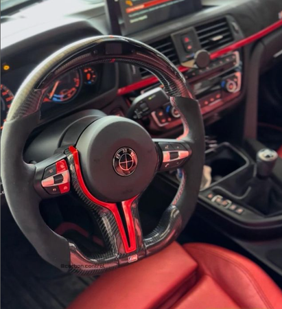 The steering wheel, featuring carbon fiber detailing and control buttons, is stationary in a car interior with red accents, digital dashboard, and central control panel. Text: "@carbon.control".