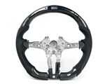 A carbon fiber steering wheel with metal interior components is displayed against a white background. Some buttons and an embedded digital display are visible on the upper section.