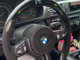 A BMW steering wheel with red stitching displays 830 rpm digitally. Surrounding dashboard shows gauges and controls. The car has a navigation screen and interior accessories on the center console.
