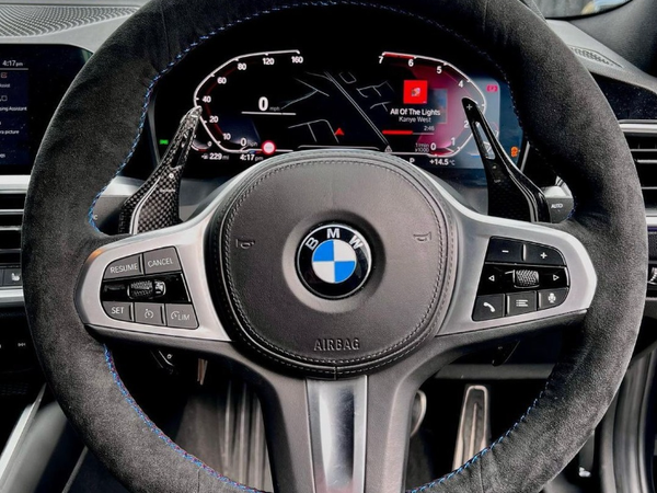A BMW steering wheel features control buttons and paddle shifters. The digital dashboard displays "All of the Lights" by Kanye West, speedometer, and various indicators. A car interior surrounds it.
