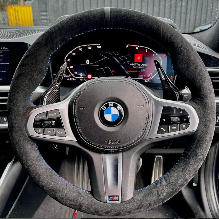 A BMW steering wheel features control buttons and paddle shifters. The digital dashboard displays "All of the Lights" by Kanye West, speedometer, and various indicators. A car interior surrounds it.