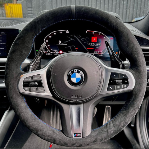 A BMW steering wheel features control buttons and paddle shifters. The digital dashboard displays "All of the Lights" by Kanye West, speedometer, and various indicators. A car interior surrounds it.